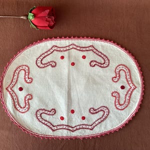 May include: Oval, cream-coloured embroidered doily with red decorative stitching and a scalloped edge. The doily features a symmetrical design with curved patterns and red dots. A red rose with a green stem is placed on the top left.