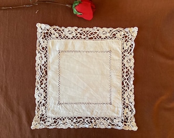 May include: A square, off-white linen doily with a delicate, openwork lace border. The center features a stitched square design. A red rose rests in the upper left corner against a brown background. This vintage textile adds a touch of elegance.