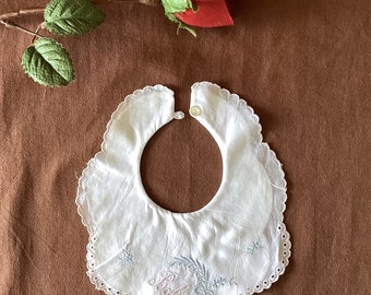 May include: White baby bib with a scalloped edge and embroidered floral details. The bib features the word "Baby" in pink script and is secured with a button closure. A red rose and stem are in the background.