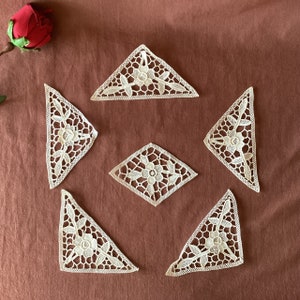 May include: Six off-white lace doilies in various shapes, including triangles and a diamond, are arranged on a brown surface. Each doily features a floral design with intricate details. A red rose is visible in the upper left corner.
