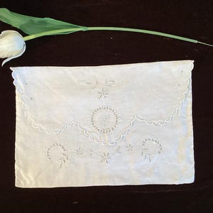 以下が含まれることがあります： 白い長方形の刺繍布。スカラップエッジがあり、花柄と円形のデザインが特徴で、中央にイニシャルがあります。白いチューリップと緑の茎が左上にあり、濃い茶色の表面に置かれています。