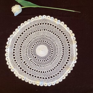 May include: A white, circular, crocheted doily with a scalloped edge. The doily features a detailed, openwork design with concentric circles. A white tulip and green stem are visible in the upper left corner.
