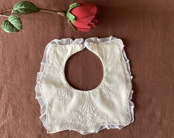 May include: Cream-colored vintage bib with delicate embroidered floral details and a scalloped lace trim. The bib is centered with a large circular opening. A red rose with green leaves rests on a brown background.
