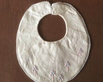 May include: A white, round baby bib with a scalloped edge and a neck opening. The bib is embroidered with small pink and blue floral designs. The bib is made of a soft, sheer fabric.