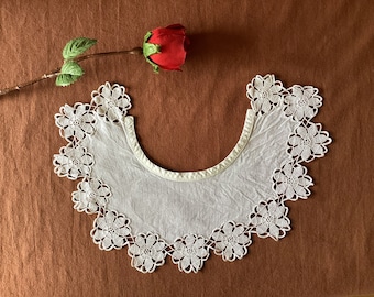May include: A white, lace collar with a floral design. The collar is crescent-shaped and edged with delicate, openwork flowers. A red rose with green leaves is in the background.
