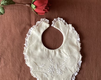 May include: Ivory-colored baby bib with embroidered floral design. The bib features a ruffled lace trim around the edges. A red rose is placed near the bib.