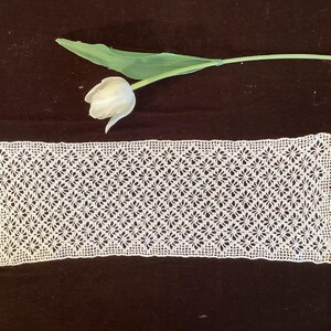 May include: A rectangular, white, open-weave lace doily with a floral pattern. A white tulip and green stem are in the background, set against a dark brown surface. The doily's intricate design is visible.