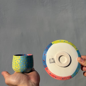 Może przedstawiać: Ręcznie robiony zestaw filiżanki i spodka ceramicznego. Filiżanka jest niebiesko-żółta w kropki. Spodek ma kolorową obwódkę. Filiżanka jest trzymana w dłoni. Spodek ma napis "E9 CERAMIC".
