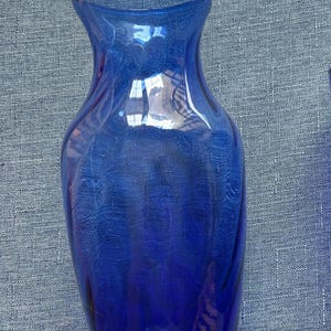 May include: A cobalt blue glass vase with a flared rim and a tapered neck. The body of the vase features a subtle, vertical ribbed texture that catches the light, creating a translucent effect throughout the deep blue glass material.