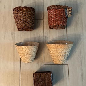 May include: Five woven planters in various styles. Two are dark brown, two are light tan, and one is a dark brown square. The planters are made of natural materials and are suitable for indoor or outdoor use.
