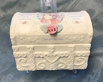Könnte beinhalten: Eine kleine, cremefarbene dekorative Truhe mit einem Klappdeckel. Die Truhe hat florale und Schnörkeldetails, einen rosa Rosenakzent und den Text "Cherish The Day". Der Deckel hat eine gemalte Illustration und Schrift. Die Truhe wird auf einem transparenten Ständer ausgestellt.
