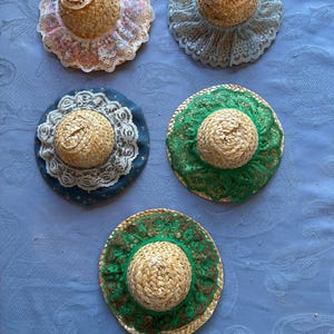 May include: Five miniature straw hats with decorative lace brims. The hats feature a woven straw crown and a wide brim adorned with lace in various colors, including pink, blue, and green. The hats are arranged on a blue surface.