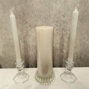 May include: Two tall white taper candles standing in clear glass candle holders with octagonal bases, positioned on either side of a thick white pillar candle resting on a clear glass plate with a scalloped, beaded edge design.