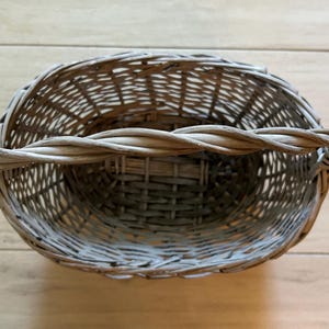May include: A gray woven wicker basket with a twisted handle. The basket has an oval shape and is made of natural materials. The handle is thick and sturdy, and the basket appears to be empty. The basket is sitting on a light brown wooden surface.