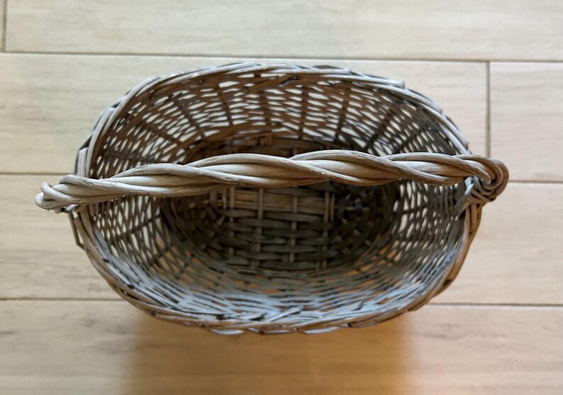 May include: A gray woven wicker basket with a twisted handle. The basket has an oval shape and is made of natural materials. The handle is thick and sturdy, and the basket appears to be empty. The basket is sitting on a light brown wooden surface.