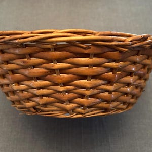 Puede incluir: Una cesta peque&ntilde;a y redonda tejida a mano con un acabado marr&oacute;n natural. El recipiente en forma de cuenco est&aacute; construido con un patr&oacute;n de tejido tradicional, mostrando hebras visibles de mimbre o junco y un borde trenzado grueso.