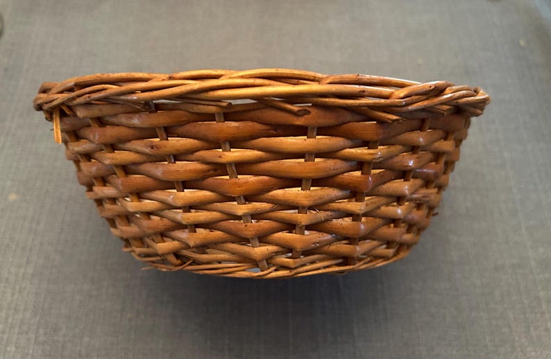 Puede incluir: Una cesta peque&ntilde;a y redonda tejida a mano con un acabado marr&oacute;n natural. El recipiente en forma de cuenco est&aacute; construido con un patr&oacute;n de tejido tradicional, mostrando hebras visibles de mimbre o junco y un borde trenzado grueso.