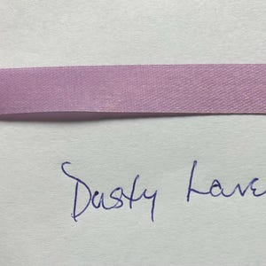 May include: A horizontal strip of smooth, dusty lavender satin ribbon with a subtle sheen. Below the ribbon, the words "Dusty Lavender" are handwritten in blue ink on a plain, light-colored surface, clearly labeling the color of the ribbon.