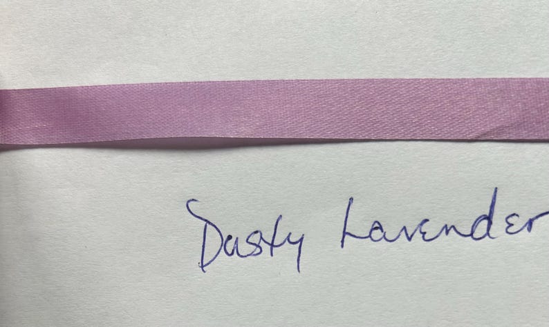 May include: A horizontal strip of smooth, dusty lavender satin ribbon with a subtle sheen. Below the ribbon, the words "Dusty Lavender" are handwritten in blue ink on a plain, light-colored surface, clearly labeling the color of the ribbon.