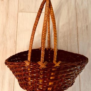 May include: A woven brown basket with a handle. The basket has a rounded shape with a wide opening and a sturdy handle made of three woven strands. The basket is made of natural materials and has a rustic appearance.