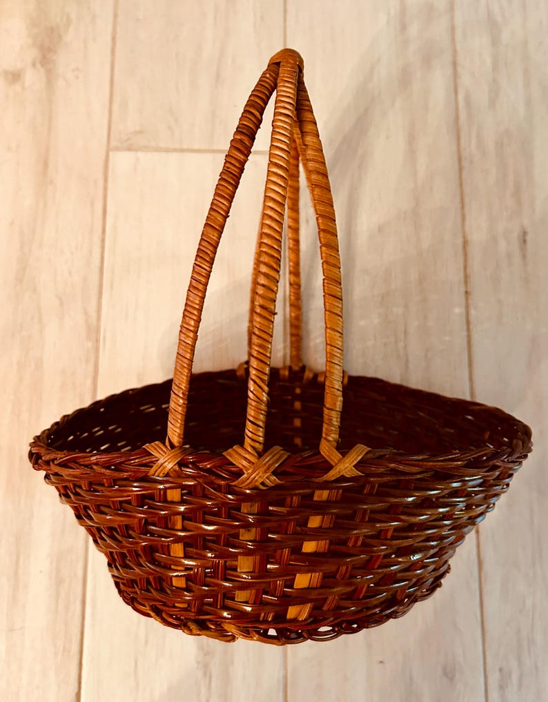 May include: A woven brown basket with a handle. The basket has a rounded shape with a wide opening and a sturdy handle made of three woven strands. The basket is made of natural materials and has a rustic appearance.