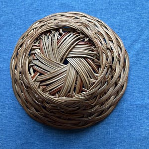 Puede incluir: Una cesta redonda y poco profunda tejida con finas ca&ntilde;as naturales de color marr&oacute;n claro. El dise&ntilde;o presenta un complejo patr&oacute;n circular superpuesto que irradia desde una abertura central, creando una estructura texturizada de tejido abierto.