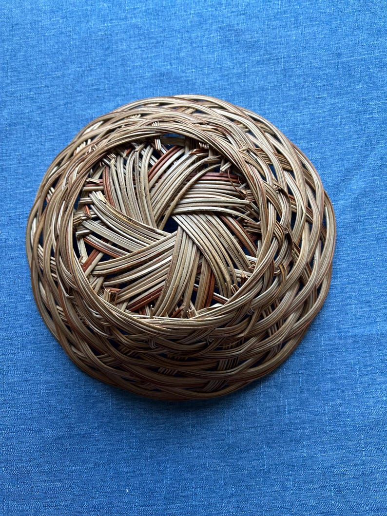 Puede incluir: Una cesta redonda y poco profunda tejida con finas ca&ntilde;as naturales de color marr&oacute;n claro. El dise&ntilde;o presenta un complejo patr&oacute;n circular superpuesto que irradia desde una abertura central, creando una estructura texturizada de tejido abierto.