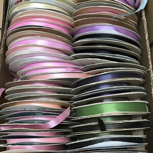 May include: A large collection of ribbon spools organized in a cardboard box. The spools feature a variety of colors including shades of pink, blue, green, purple, yellow, and black. Each spool is made of cardboard and holds a single color of ribbon.