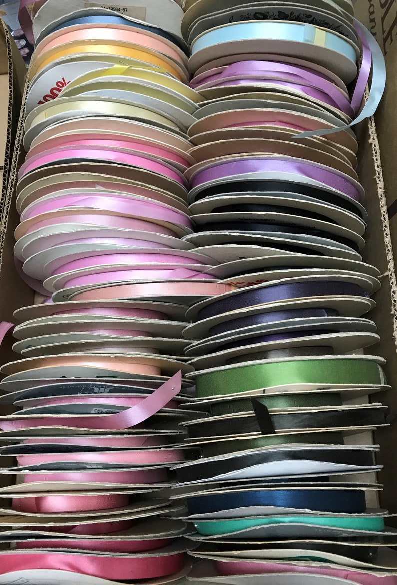 May include: A large collection of ribbon spools organized in a cardboard box. The spools feature a variety of colors including shades of pink, blue, green, purple, yellow, and black. Each spool is made of cardboard and holds a single color of ribbon.