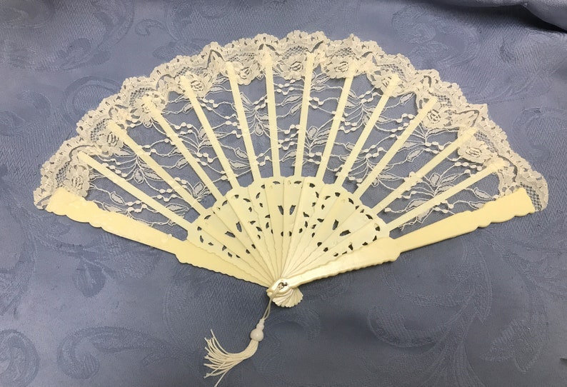May include: A white lace fan with a carved ivory handle. The fan is closed and has a tassel hanging from the bottom.