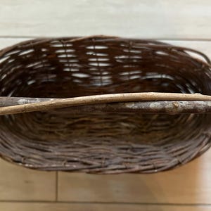 May include: Oval-shaped woven basket with a natural brown color. A light-colored wooden handle is attached across the top. The basket is made of woven twigs and branches, and is empty.