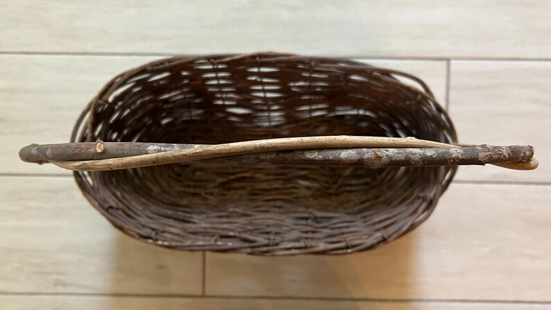 May include: Oval-shaped woven basket with a natural brown color. A light-colored wooden handle is attached across the top. The basket is made of woven twigs and branches, and is empty.