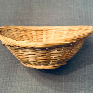May include: A small, woven basket made of light brown wicker. The basket has a rounded shape with a slightly flared rim. The weave is tight and even, creating a sturdy container. The basket is empty and sits on a gray surface.