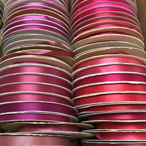 May include: A collection of numerous circular spools of satin ribbon arranged in two vertical columns. The ribbons feature a variety of shades including bright pink, magenta, deep red, burgundy, and soft mauve, all with a smooth, reflective finish.