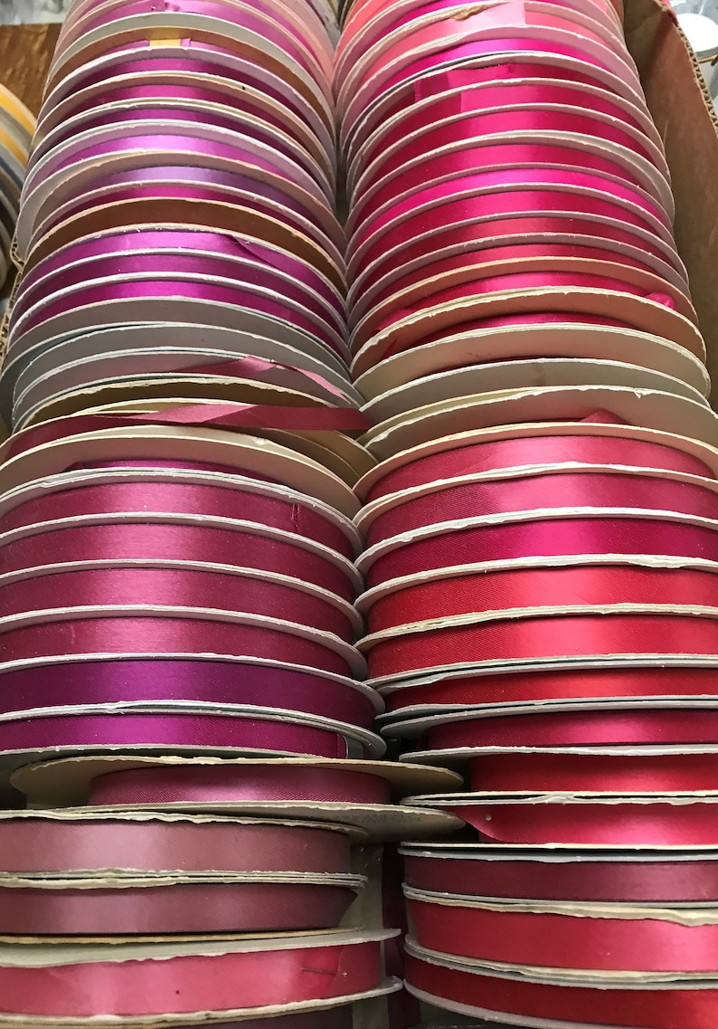 May include: A collection of numerous circular spools of satin ribbon arranged in two vertical columns. The ribbons feature a variety of shades including bright pink, magenta, deep red, burgundy, and soft mauve, all with a smooth, reflective finish.
