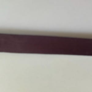 May include: A dark brown ribbon with a fine, horizontal ribbed texture. The ribbon is positioned horizontally across the frame, displaying a consistent, solid dark brown color throughout its length and a subtle, woven fabric appearance.
