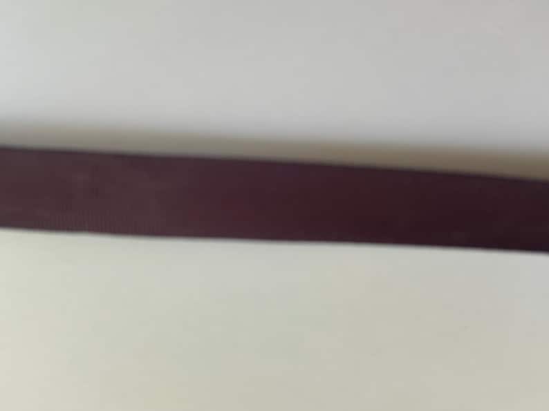 May include: A dark brown ribbon with a fine, horizontal ribbed texture. The ribbon is positioned horizontally across the frame, displaying a consistent, solid dark brown color throughout its length and a subtle, woven fabric appearance.