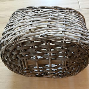 May include: A woven basket with a square shape. The basket is made of light brown and gray wicker, with a tightly woven pattern. The basket is sitting on a light brown wooden surface.