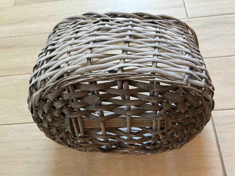 May include: A woven basket with a square shape. The basket is made of light brown and gray wicker, with a tightly woven pattern. The basket is sitting on a light brown wooden surface.