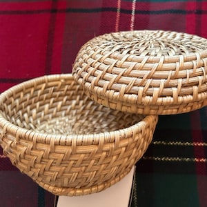 May include: A woven wicker basket with a lid. The basket is light brown and has a round shape. The lid is slightly domed and fits snugly on top of the basket.