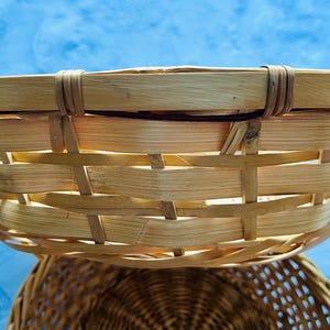 Puede incluir: Una cesta redonda y poco profunda construida con tiras de madera natural de color claro tejidas en un patr&oacute;n horizontal. El borde est&aacute; reforzado con una banda de madera lisa y gruesa asegurada por tiras verticales, creando un recipiente abierto.