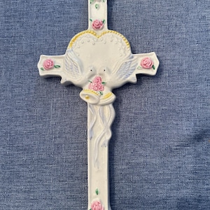 May include: White ceramic cross with a heart-shaped design and two doves holding a bell. The cross is decorated with pink roses and green leaves.