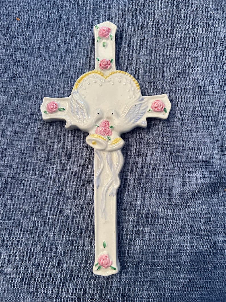 May include: White ceramic cross with a heart-shaped design and two doves holding a bell. The cross is decorated with pink roses and green leaves.
