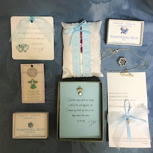 May include: A set of six blue and white themed wedding favors. The favors include a blue pin, a blue toe ring, a blue bracelet, a blue angel charm, a blue angel pin, and a blue and white satin pillow with a blue lace trim. The favors are packaged in small boxes with blue and white designs. The boxes have text that says "Something Old, Something New, Something Borrowed, Something Blue".