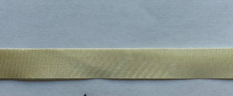 May include: A single strip of smooth, light gold-colored ribbon with a subtle sheen and a tightly woven texture. The ribbon is positioned horizontally across the frame, displaying a consistent width and a flat, uniform appearance throughout its length.