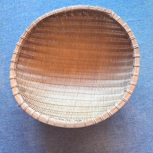 Puede incluir: Una cesta tejida redonda y poco profunda de color tostado natural. La cesta est&aacute; construida con tiras verticales finas similares al bamb&uacute; y un borde circular reforzado y m&aacute;s grueso, unido a intervalos regulares con material de color claro.