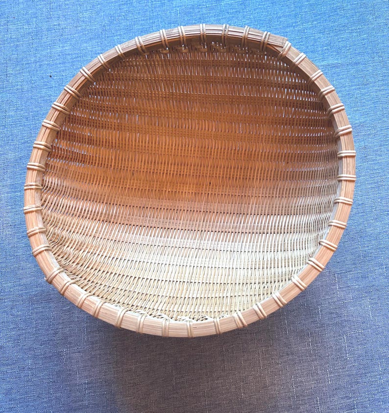 Puede incluir: Una cesta tejida redonda y poco profunda de color tostado natural. La cesta est&aacute; construida con tiras verticales finas similares al bamb&uacute; y un borde circular reforzado y m&aacute;s grueso, unido a intervalos regulares con material de color claro.