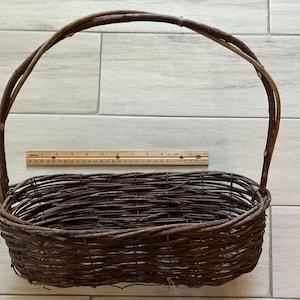 May include: A brown woven basket with a handle. The basket is oval-shaped and made of intertwined twigs. A wooden ruler is placed next to the basket. The basket's dimensions are approximately 14 inches long and 8 inches wide.