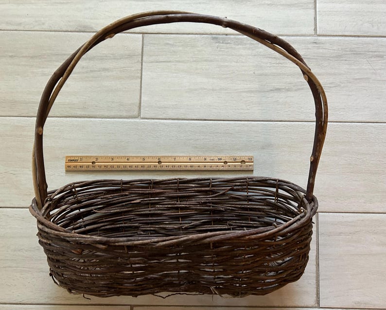May include: A brown woven basket with a handle. The basket is oval-shaped and made of intertwined twigs. A wooden ruler is placed next to the basket. The basket's dimensions are approximately 14 inches long and 8 inches wide.