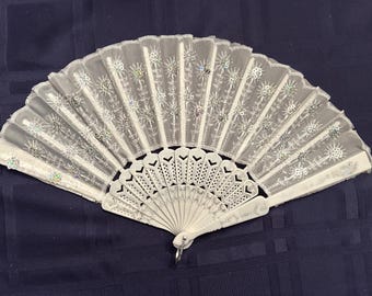 May include: A white folding hand fan featuring a sheer fabric leaf adorned with silver embroidered floral patterns and iridescent sequins. The handle and ribs are made of white plastic with intricate cut-out heart and geometric designs.